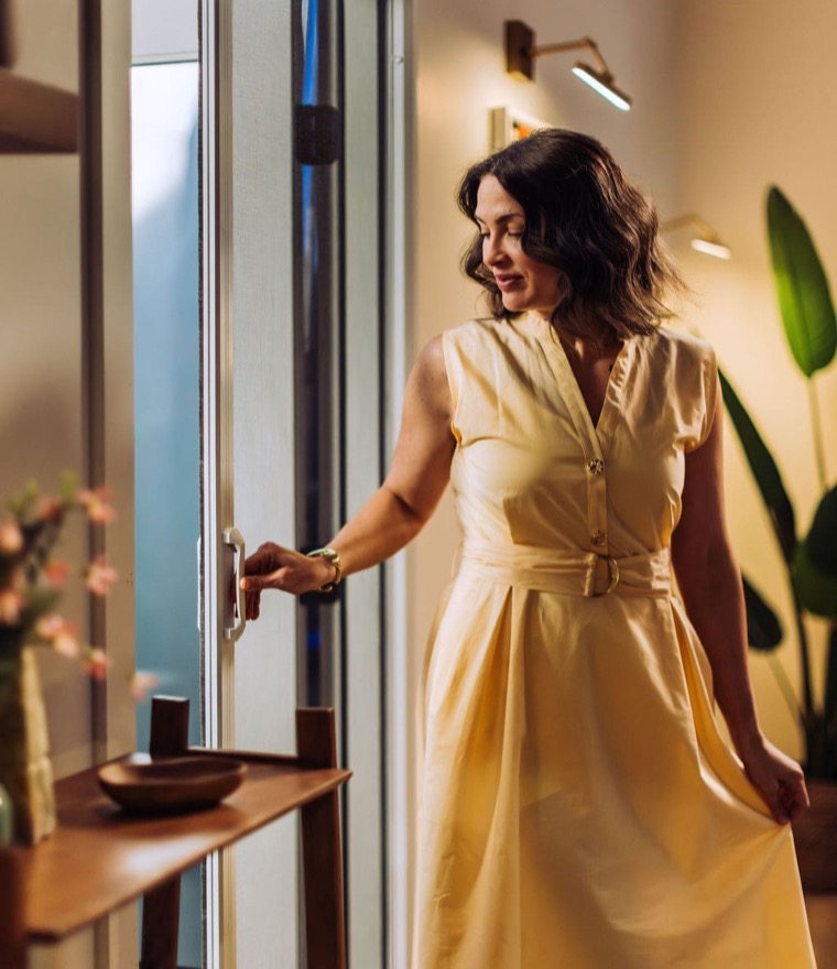 Woman in a flowing yellow dress opening a retractable screen door in a warmly lit room