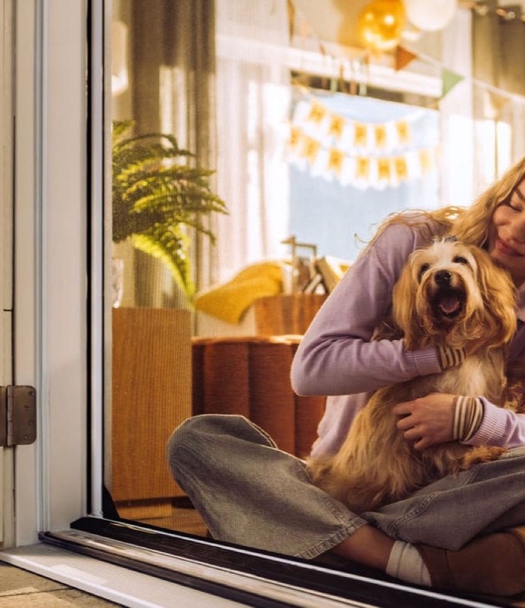 Woman sitting cross-legged on the floor hugging a small dog beside an open retractable screen door, with a party banner in the background