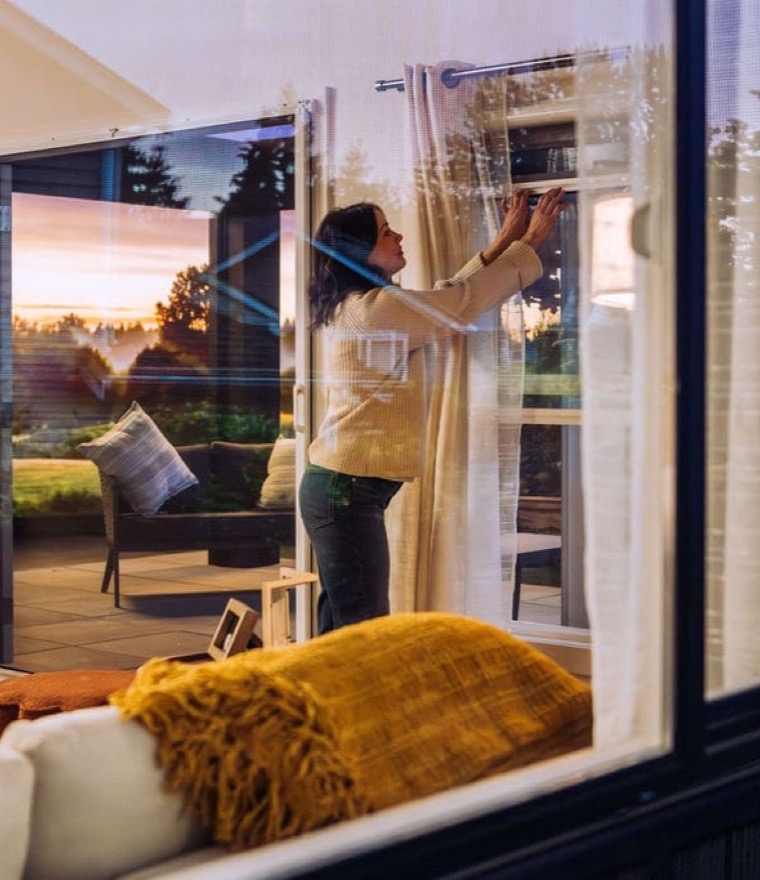 Woman lowering a retractable window screen at dusk with a sunset and patio visible beyond